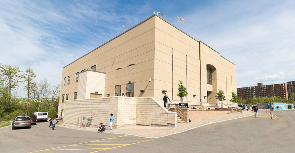 Exterior photo of a large rectangular beige coloured building and an apartment building in the background.